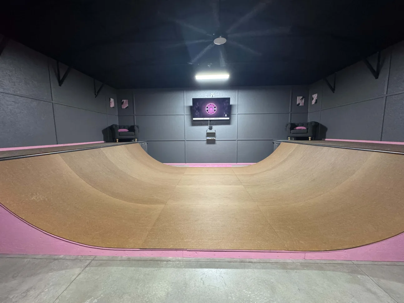 The Wilson Wheels indoor wooden mini ramp with pink trim, dark walls, and a mounted screen